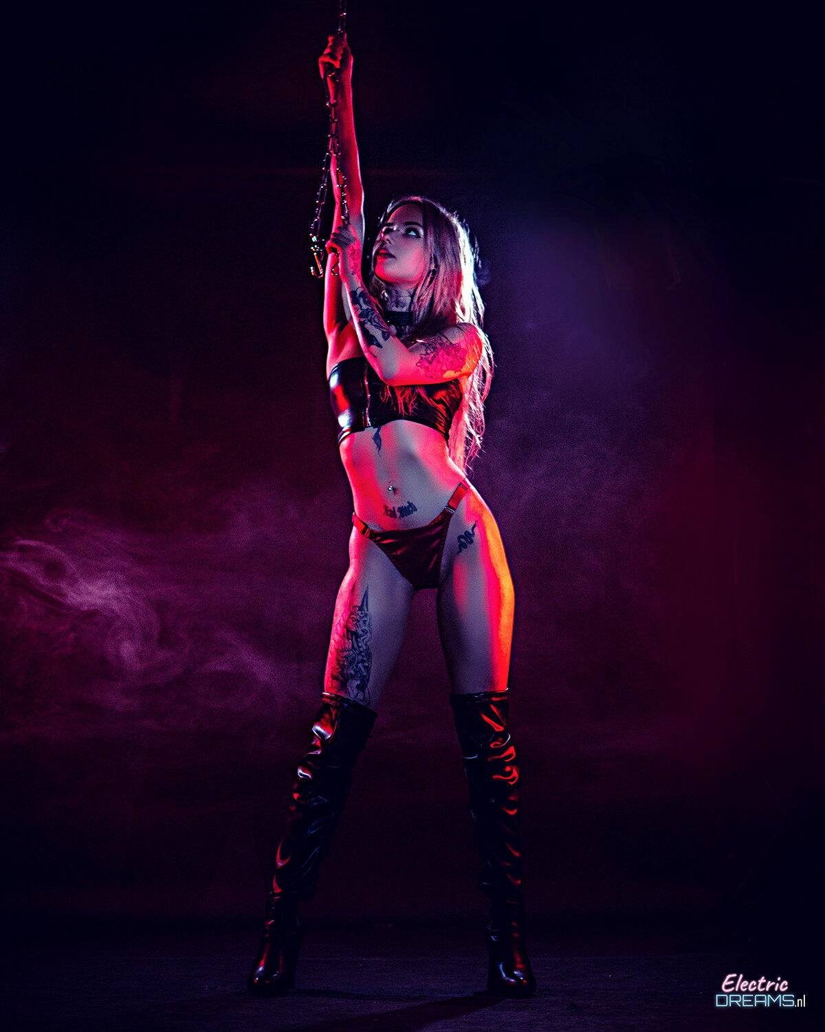 The image shows a woman in a red bikini and thigh high boots standing on a stage, holding an object in her hand. The background is dark and there is a watermark on the image.