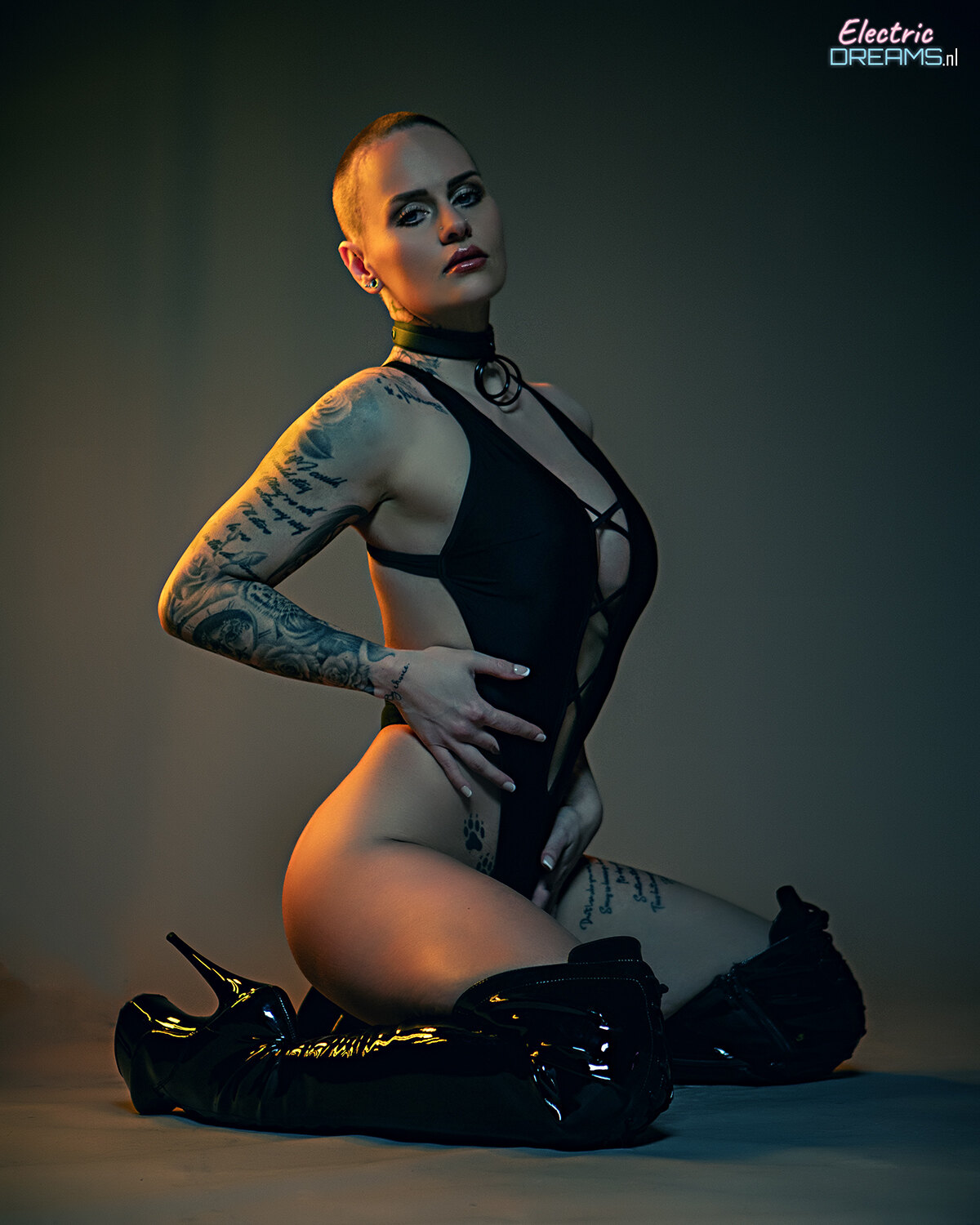 The image shows a woman in a black bodysuit sitting on the ground with tattoos on her arms and legs. She is wearing high heels, giving her a stylish and confident look.