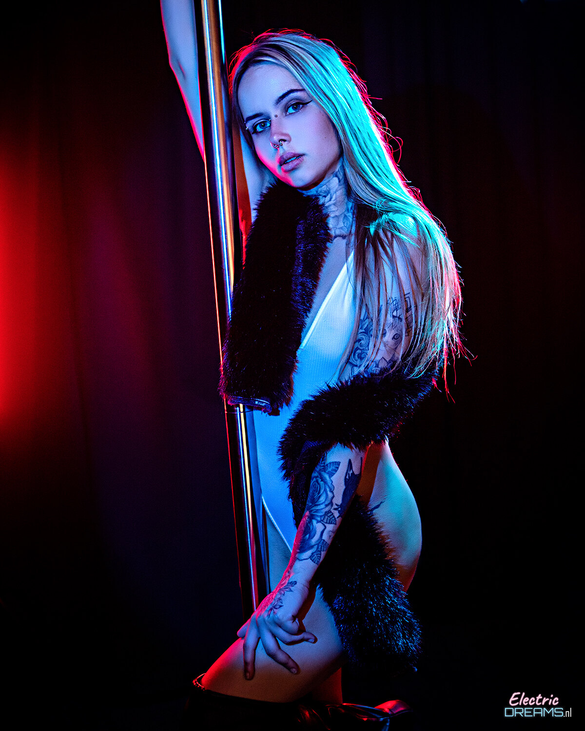 The image shows a woman in a white dress standing on a pole in a dark background, illuminated by a red light.