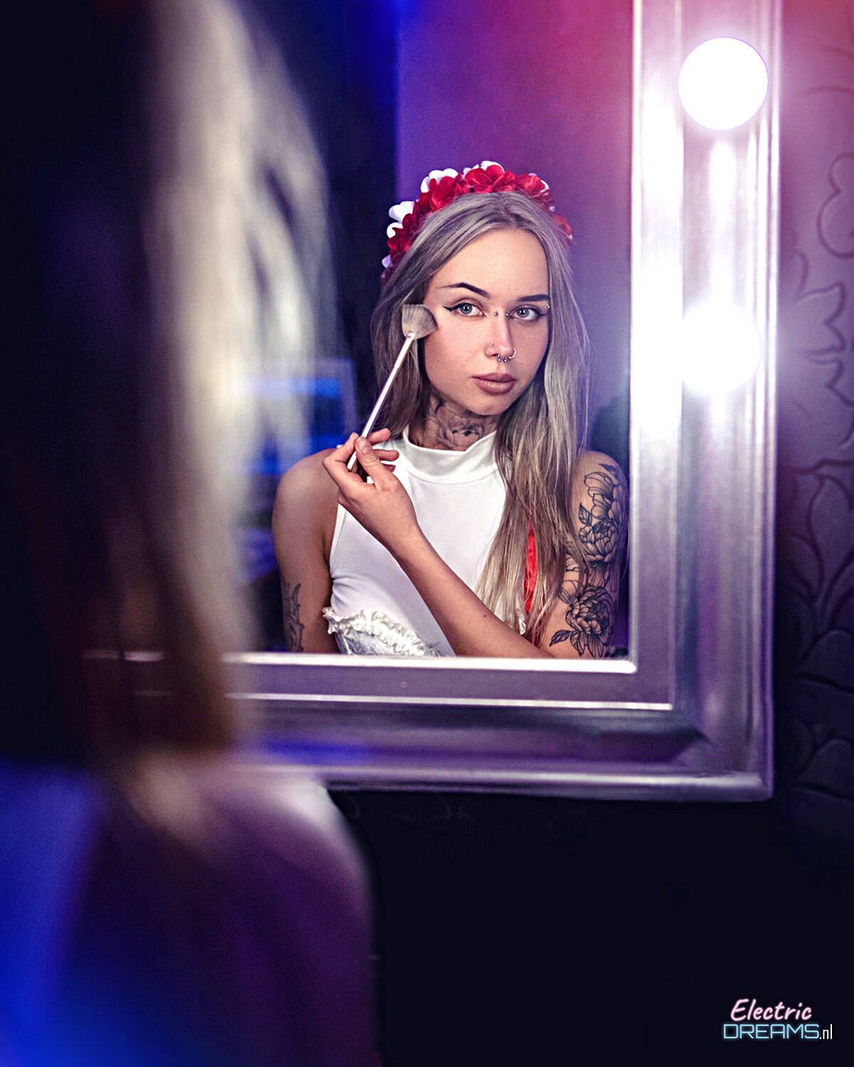 The image shows a woman in a white dress with a flower crown on her head, holding a makeup brush in her hand and looking at her reflection in the mirror. There is a light illuminating her face and a watermark in the bottom right corner of the image.