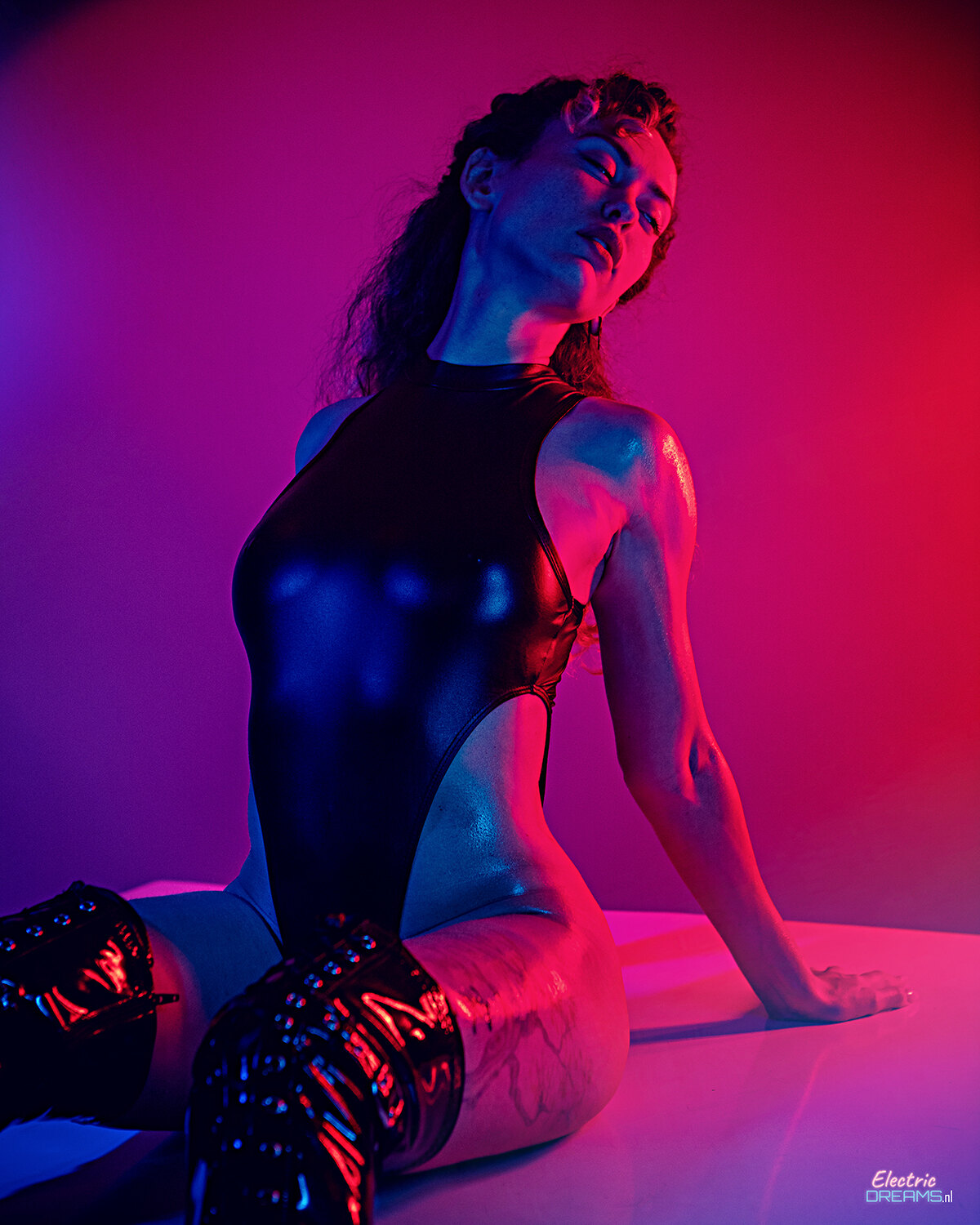 The image shows a woman in a black bodysuit sitting on a white surface with a pink and red background. She has a human face and is wearing footwear.