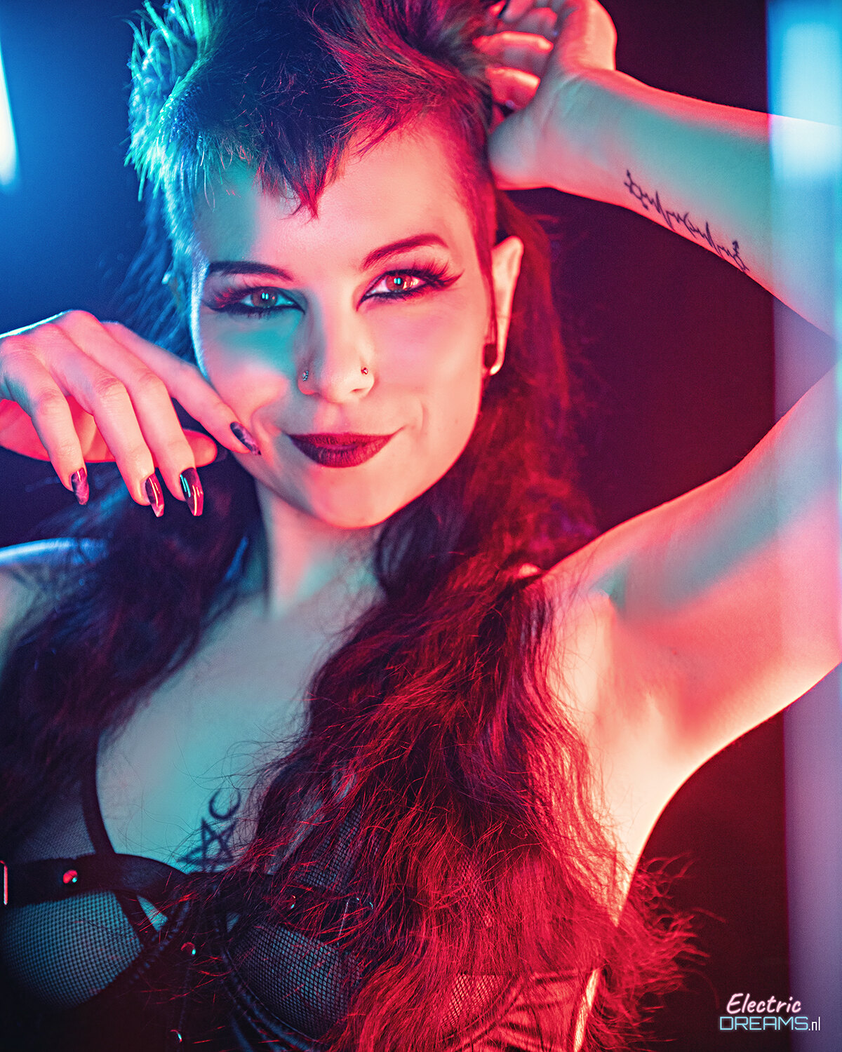 The image shows a woman with long red hair and a tattoo on her arm, standing in front of a wall with lights in the background and text at the bottom.