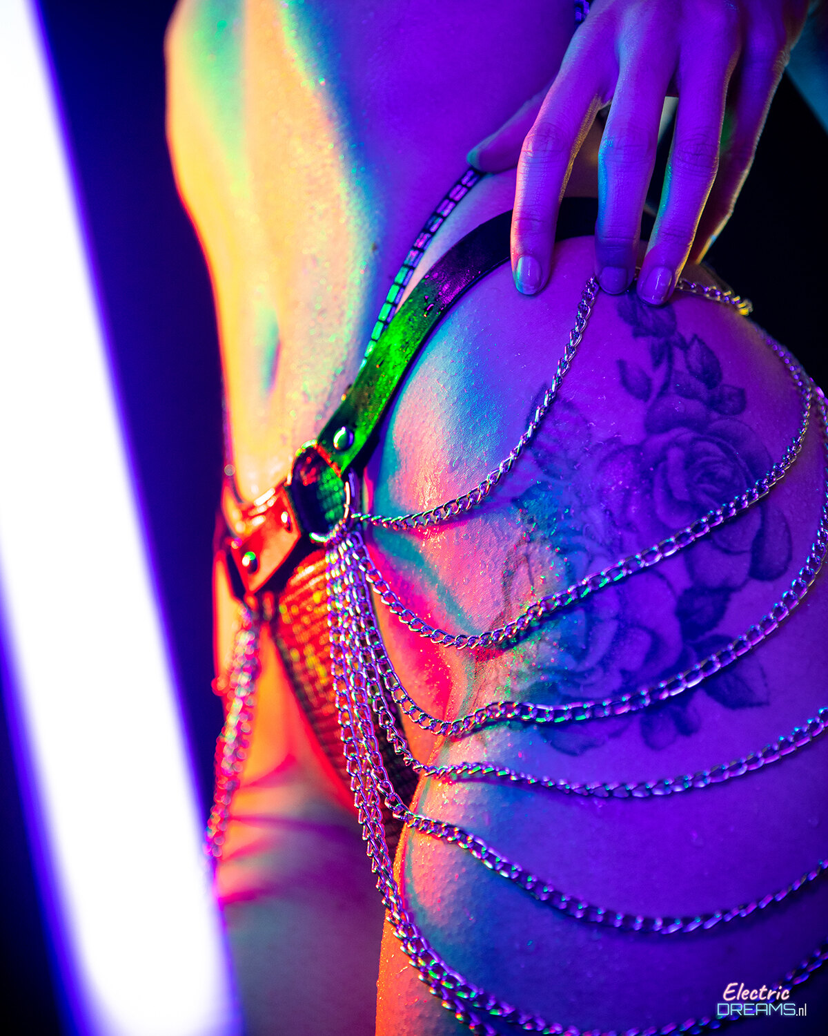 The image shows a woman with a tattoo on her back and a chain around her waist, illuminated by a light on the left side.