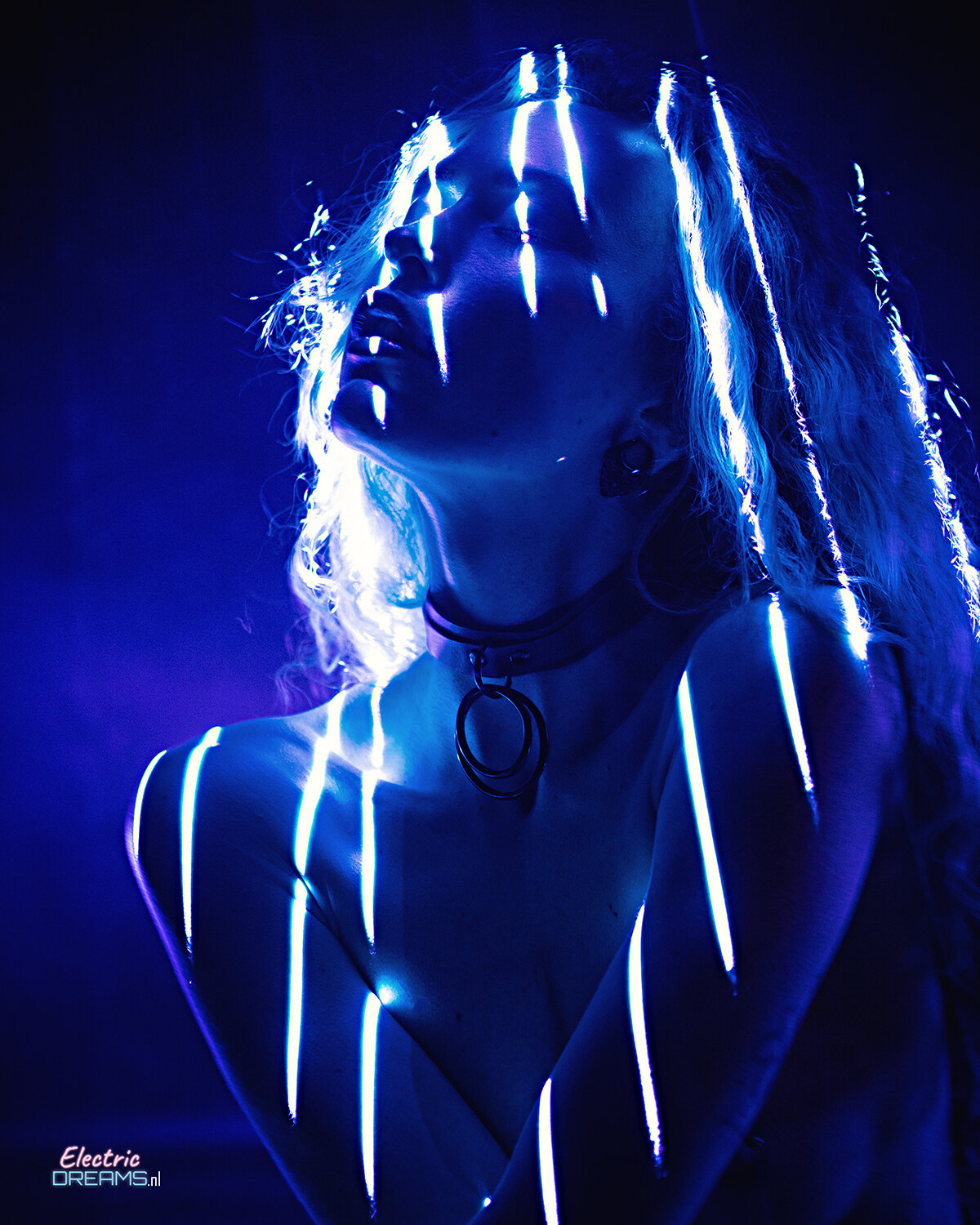 The image shows a woman with long blonde hair and blue lights on her body, illuminated against a dark background with a watermark.