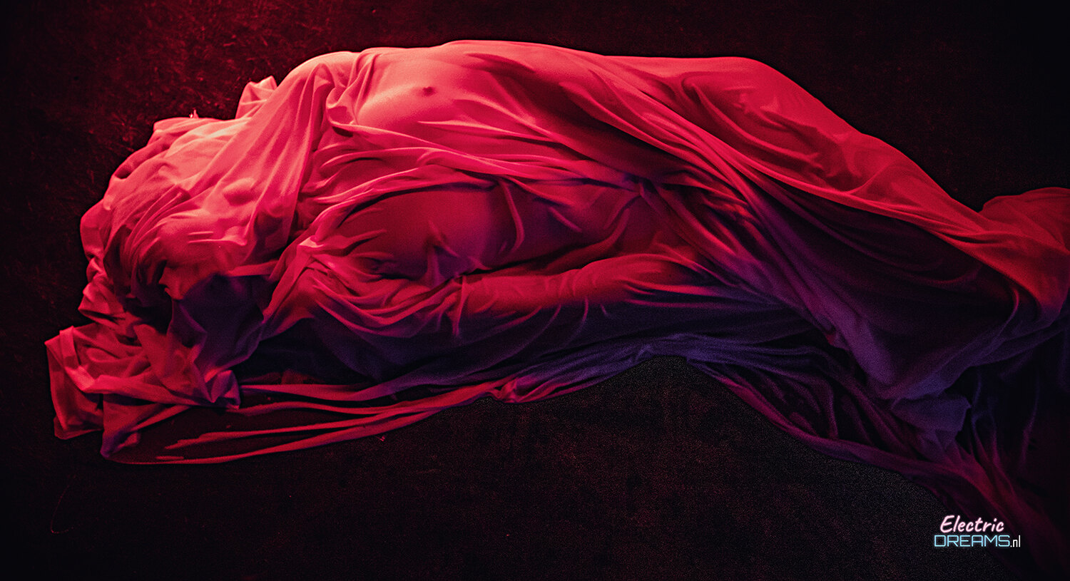 The image shows a woman in a red dress laying on a black surface, with a watermark in the bottom right corner. Her dress is a vibrant red color, and her hair is pulled back in a neat bun. She appears to be sleeping peacefully, her eyes closed and her head