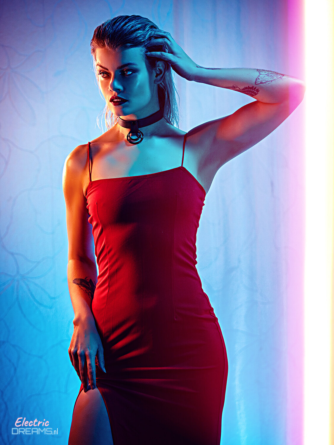 The image shows a woman in a red dress posing for a picture. She has a confident and poised stance, her hair is styled in a sleek bob, and her makeup is subtle and natural. She is wearing a pair of black heels and her eyes are looking directly at the came