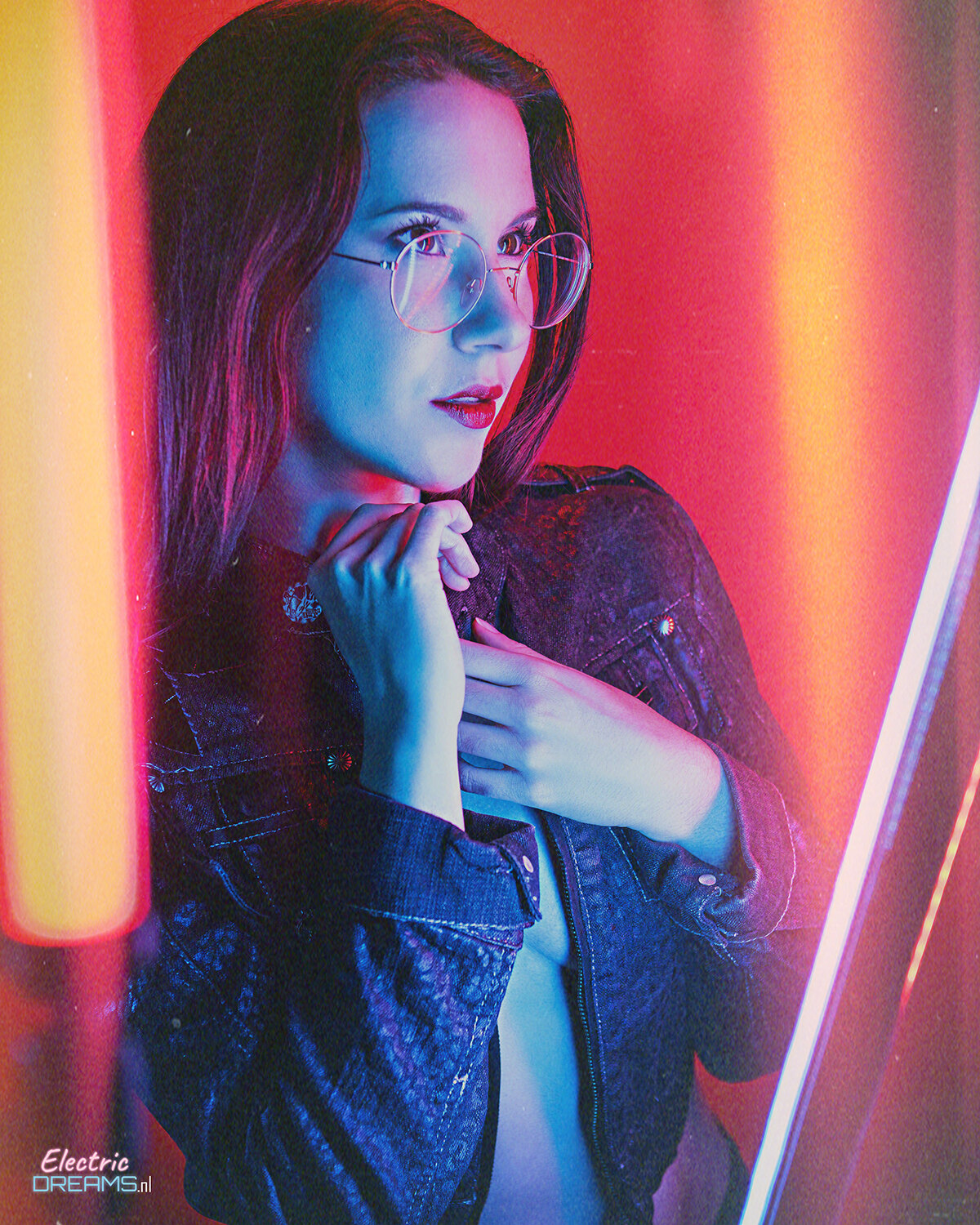 The image shows a woman wearing glasses and a denim jacket standing in front of a neon sign. She has a confident and determined expression on her face, and her hair is pulled back in a neat bun. The neon sign behind her is bright and vibrant, adding to th