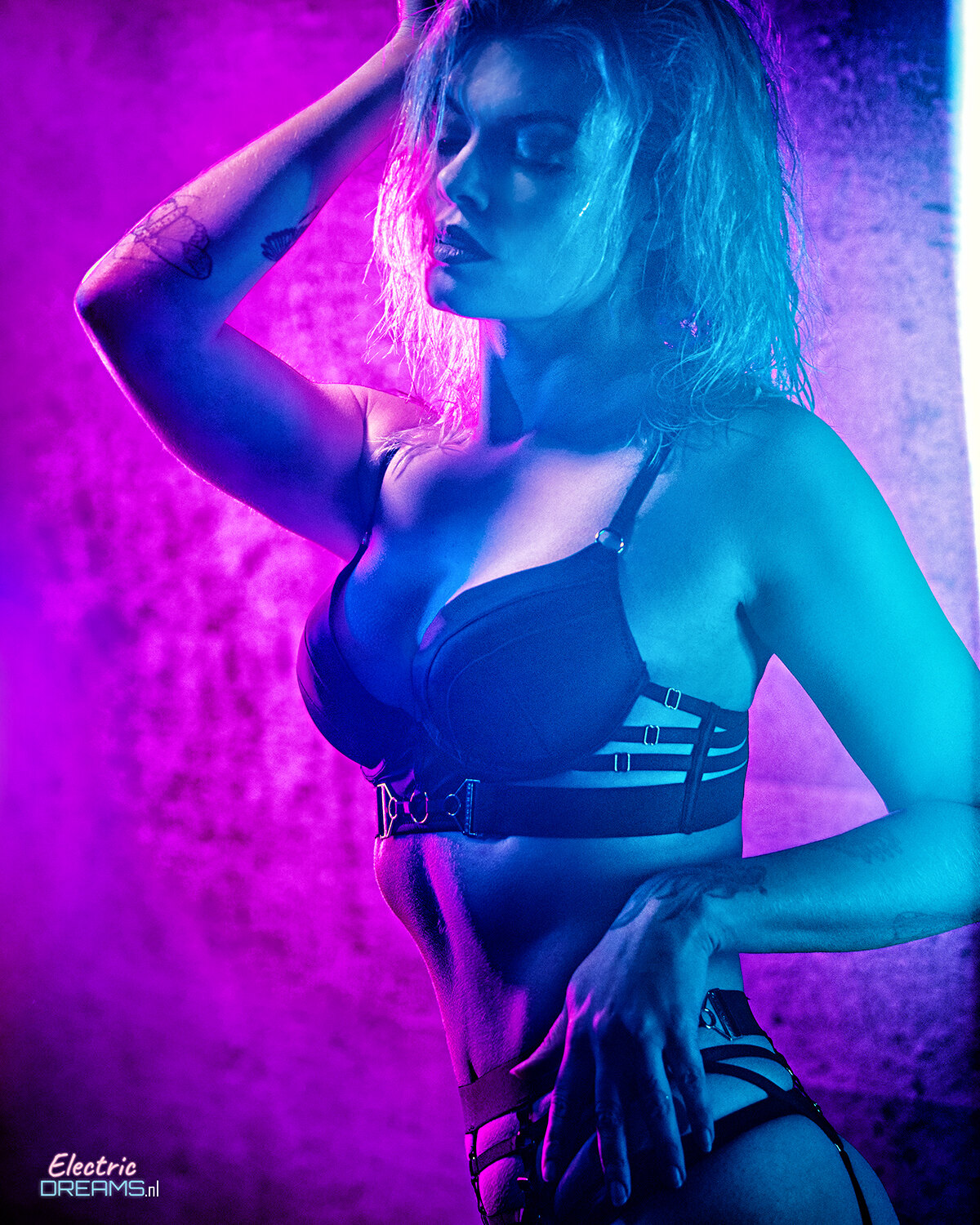 The image shows a woman in a black lingerie posing for a picture in front of a wall with text at the bottom left corner.
