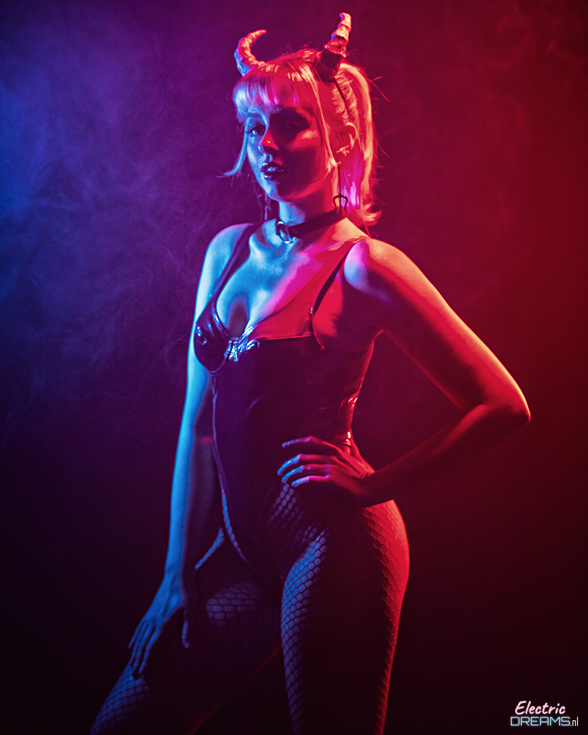 The image shows a woman in a black bodysuit with horns on her head, standing against a dark background with smoke in the background.
