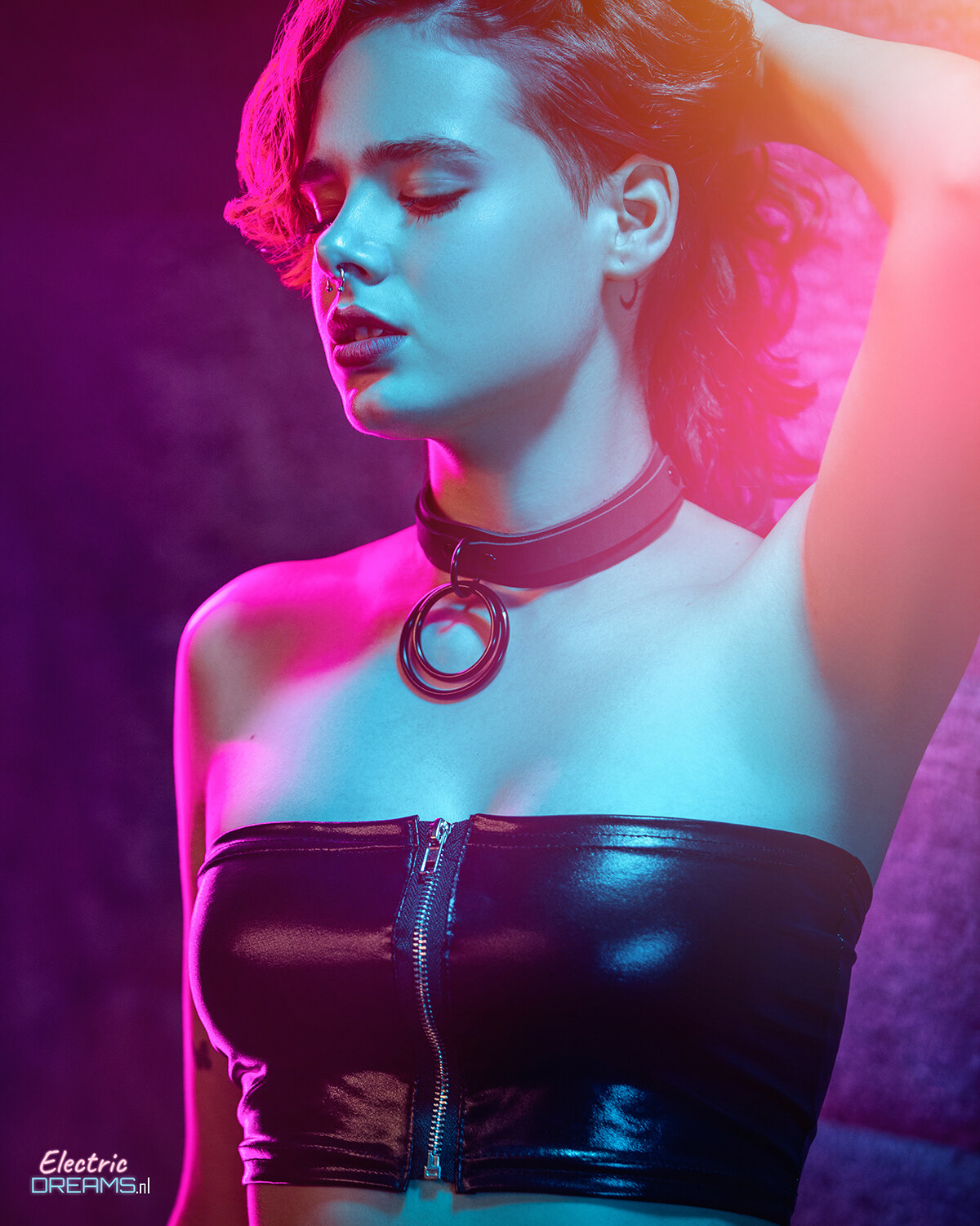 The image shows a woman wearing a black leather top and a necklace, standing in front of a wall with text at the bottom left corner.