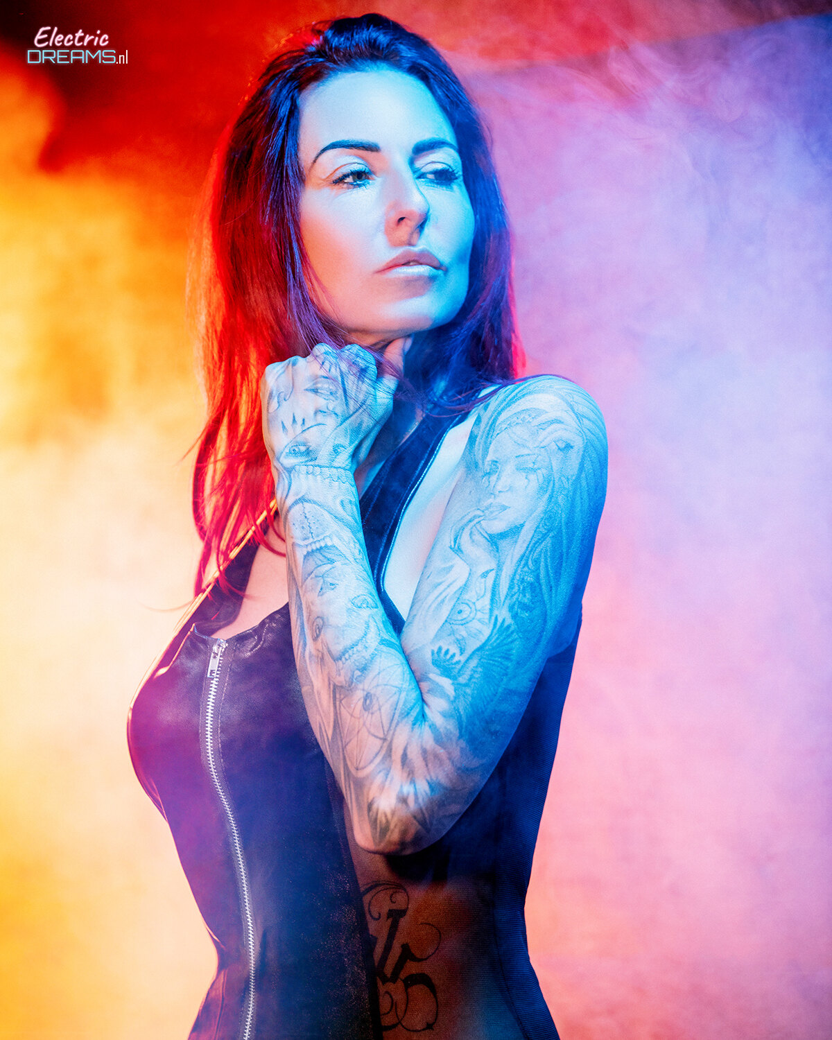 The image shows a woman in a black dress with tattoos on her arms and chest, standing in front of a colorful background with a watermark in the top left corner.