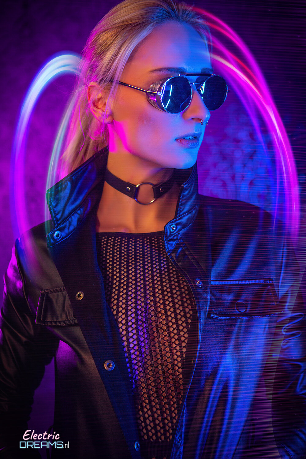 The image shows a woman wearing a black jacket and sunglasses standing in front of a neon light, with a watermark in the bottom left corner.
