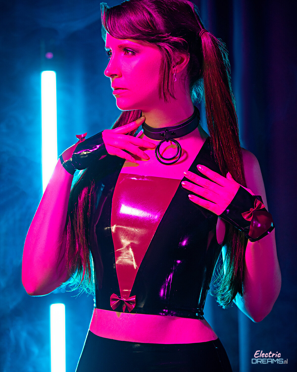 The image shows a woman in a black and pink outfit standing in front of a neon light, with a dark background and a watermark in the bottom right corner.