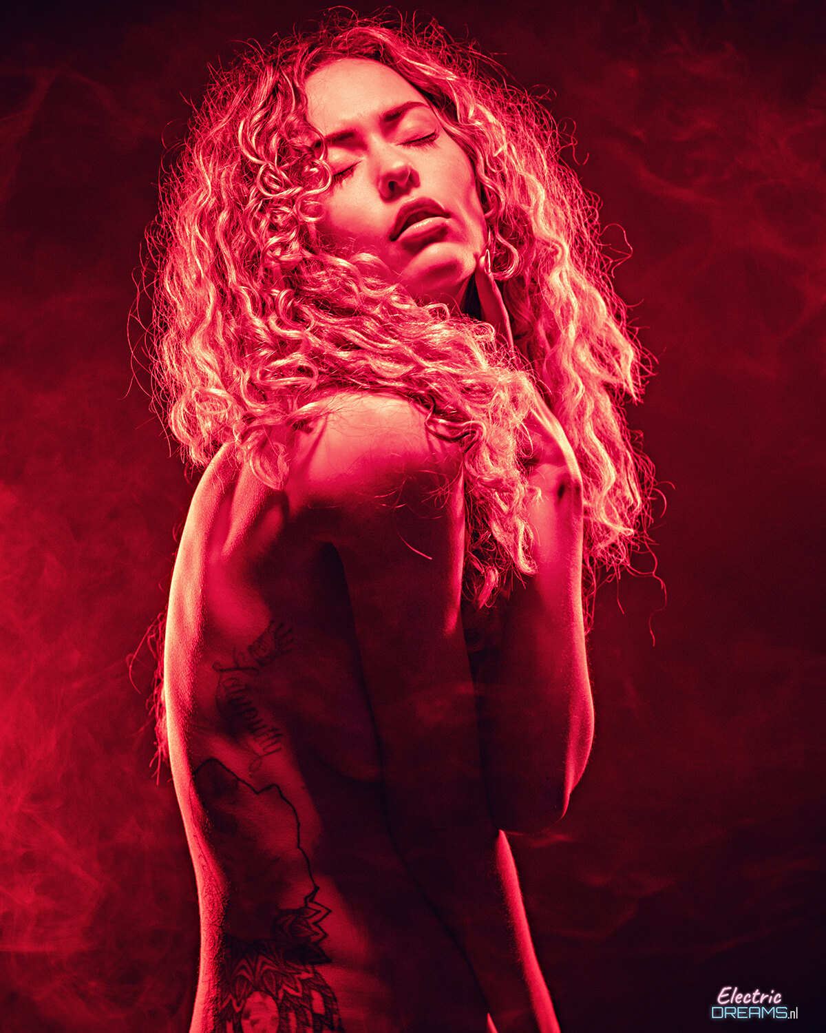 The image shows a woman with long curly hair and tattoos on her body standing in front of a red background.
