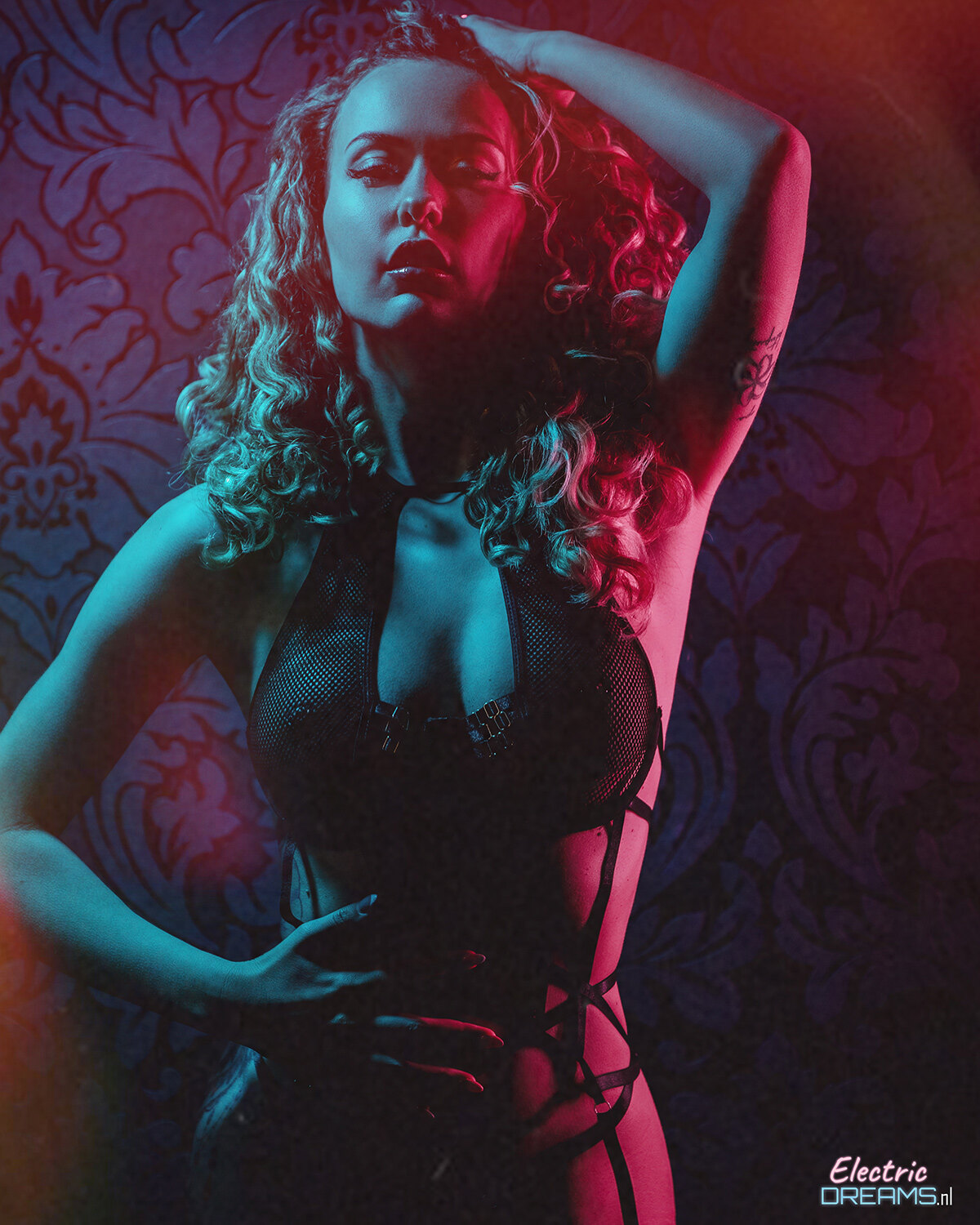 The image shows a woman in a black lingerie posing for a picture in front of a wall with a design. At the bottom right corner of the image, there is some text.