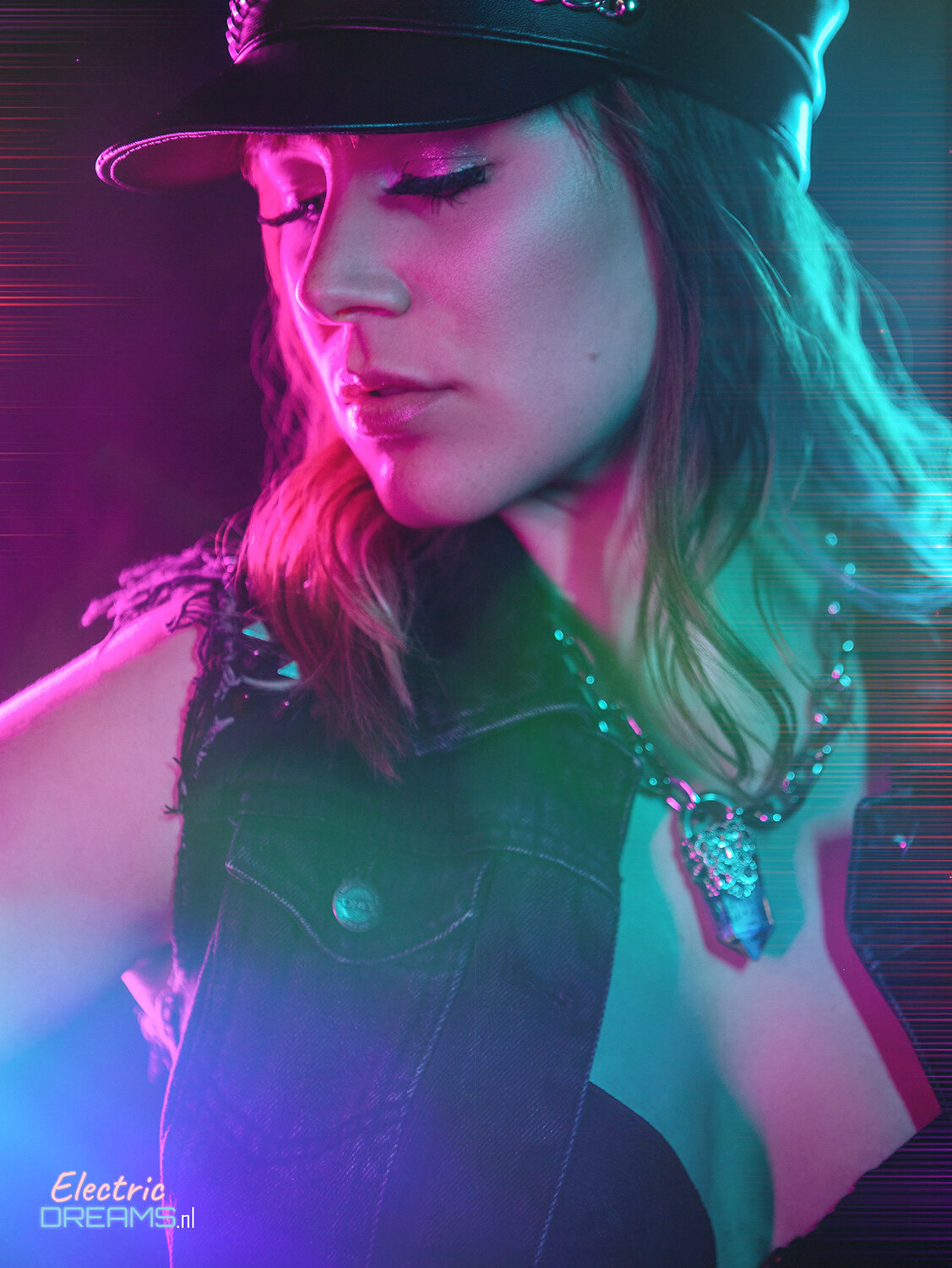The image shows a woman wearing a black leather jacket and a black hat, with a chain around her neck, standing in front of a colorful background.
