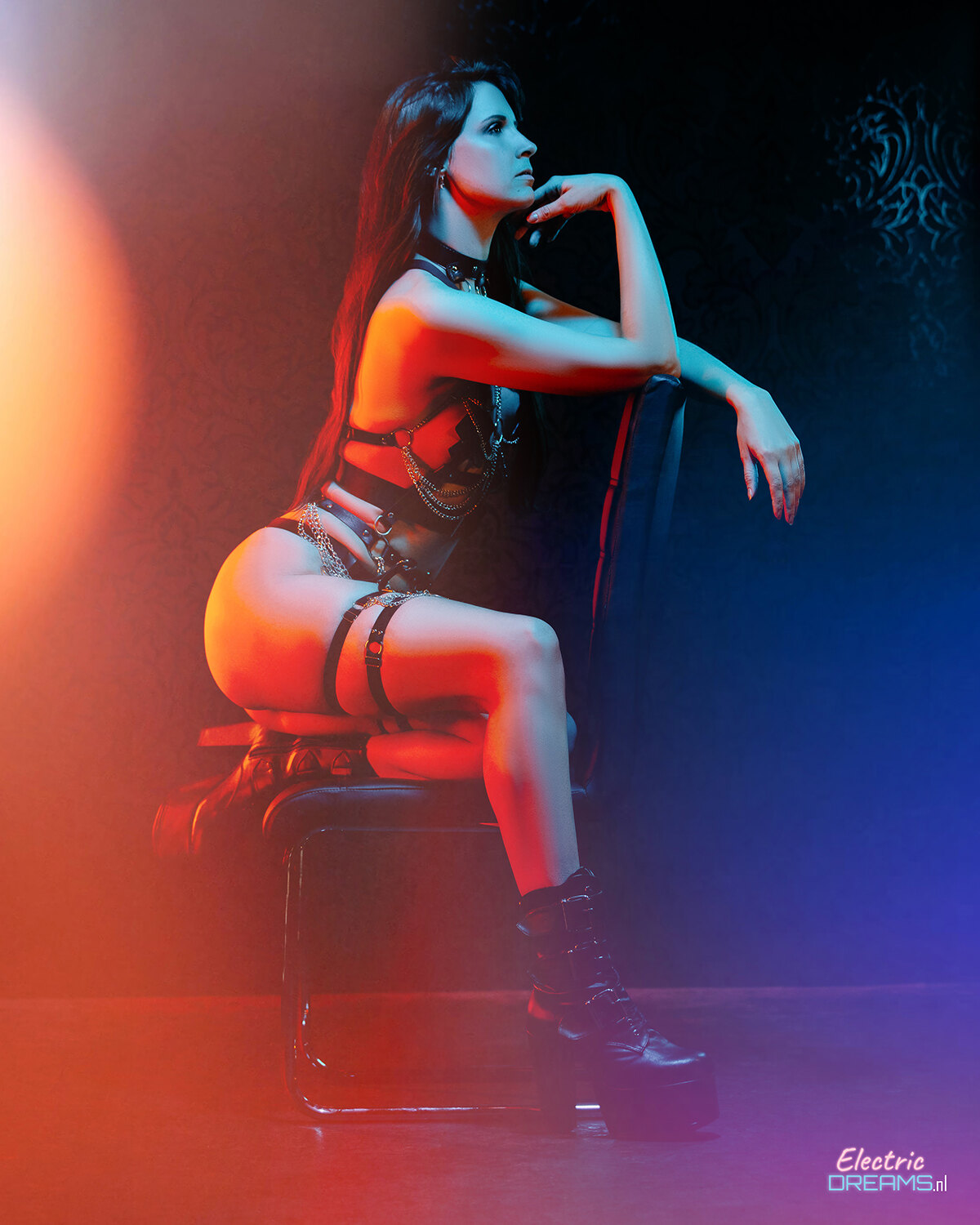 The image shows a woman sitting on top of a chair in a dark room, illuminated by a light on the left side.