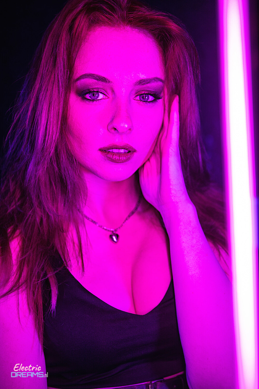 The image shows a woman in a black dress standing in front of a neon light, with a dark background and a watermark in the bottom left corner.