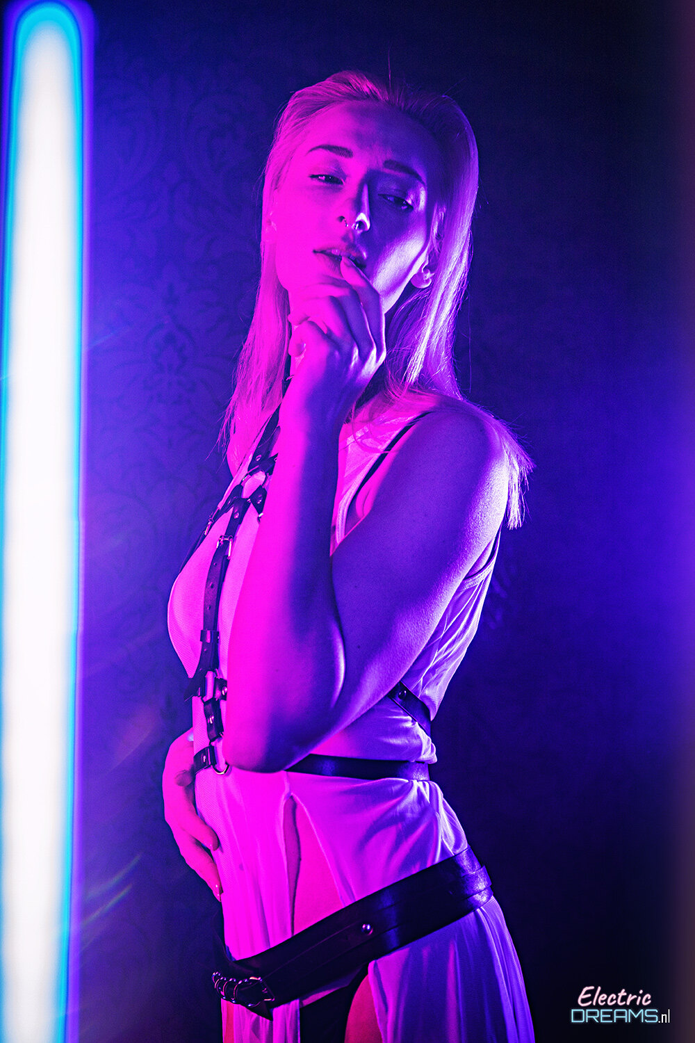 The image shows a woman in a white dress standing in front of a neon light, holding a microphone in her hand. The background is dark and there is a watermark on the image.