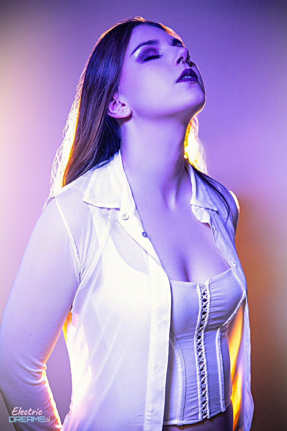 The image shows a woman in a white shirt posing for a picture. She has a confident and poised stance, her arms crossed and her head held high. Her hair is pulled back in a neat bun, and her eyes are looking directly at the camera. She is wearing a pair of