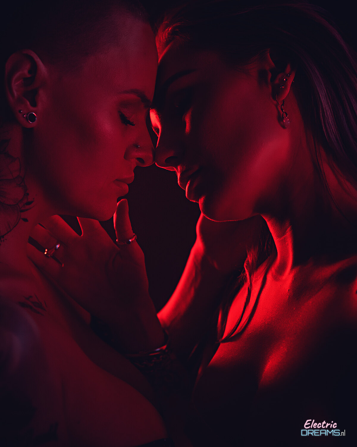 The image shows two women embracing in a dark room, their faces illuminated by a red light.
